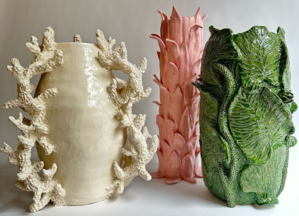 a trio of vessels, on the left is white with coral like handes, the middle is pink with scalloped leaves, and the right is green with large cabbage like leaves