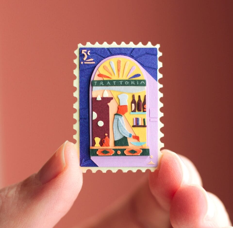 a hand holds up a small, vibrant collage in the shape of a stamp. it depicts a scene with a building and patterned designs