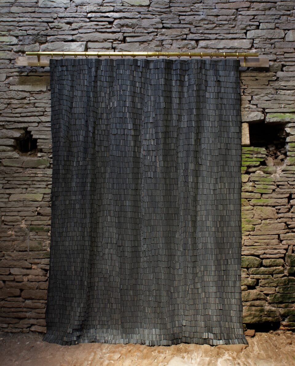 a hanging tapestry-like artwork made of tiny, dark, wooden shingles, photographed against a stone wall