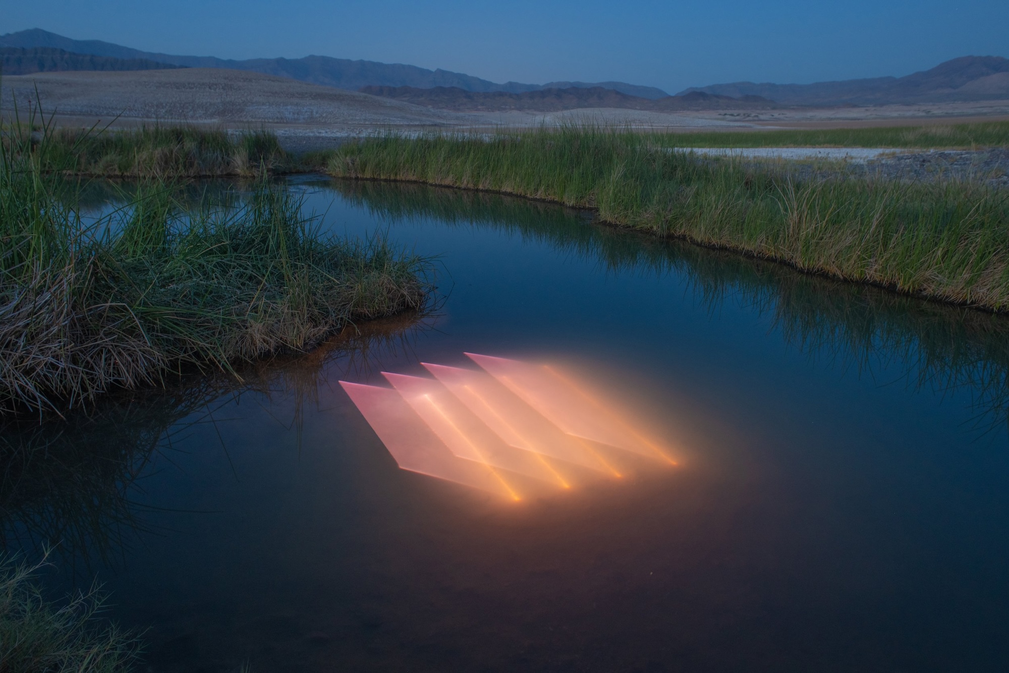 a river landscape in a desert with a peach-colored geometric light projection in the surface of the water